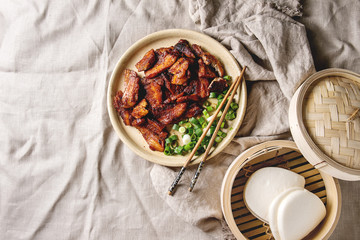 Chinese traditional dish Cantonese BBQ Pork Belly with spring onion served in ceramic plate with chopsticks, bamboo steamer and gua bao bus over linen cloth. Flat lay, space. Asian style dinner.