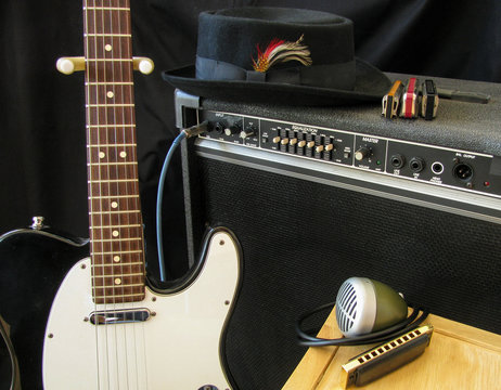 A musician’s gear on stage for performance includes black electric guitar, amplifier, blues harmonicas and microphone and pork pie hat.