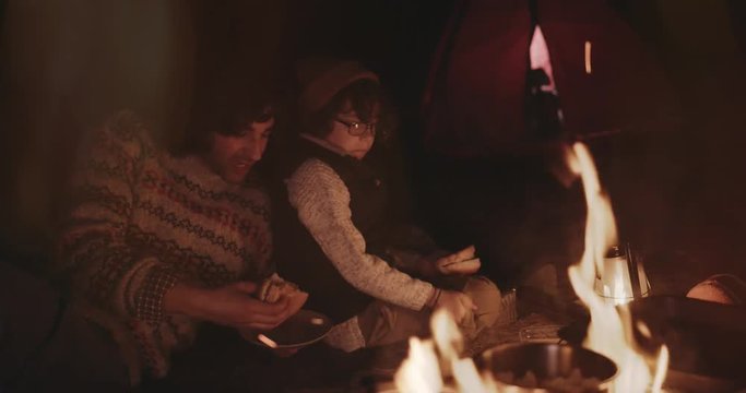 Young father and son campers eating around campfire at night