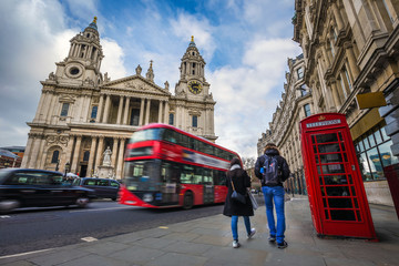 London, England - Tourist couple walking by a traditional red telephone box with red double-decker bus and black taxi on the move at background at St.Paul's Cathedral