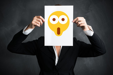 junge Geschäftsfrau hält Schild mit schockiertem Smiley vor ihr Gesicht