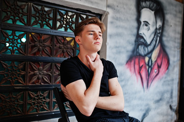 Young stylish macho boy in black jacket posed outdoor of street. Amazing model man sitting against grafiti wall of barbershop.