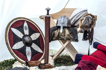 Medieval knight equipment in old sleeping tent. Metal helmet, shield, sword and clothes on chair....