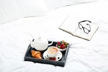 Tasty breakfast in bed on wooden tray with book and glasses
