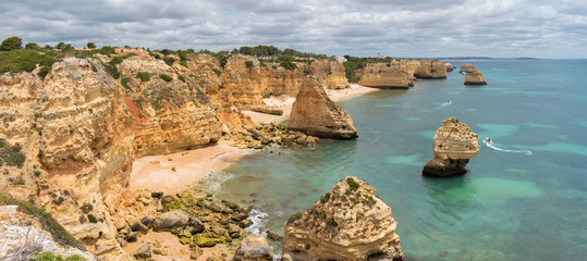 Fototapeta na wymiar Panorama of Praia da Marinha on the south coast of Portugal in the Algarve on a cloudy day. The cliffs, beach and Atlantic ocean are in view with tourists on the beach and a couple of boats in the bay