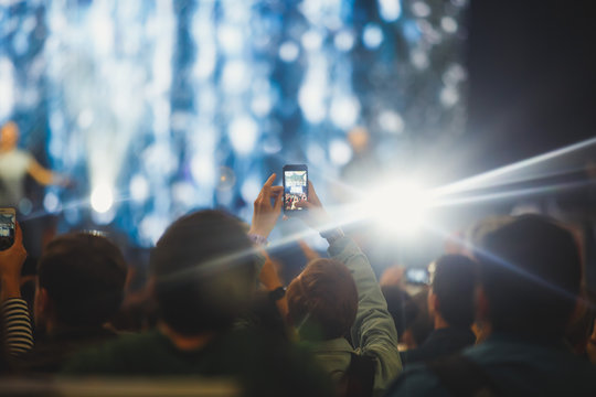 A crowded concert hall with scene stage lights, rock show performance, with people silhouette and hand holding smartphone