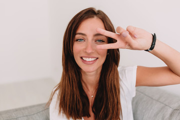 Fototapeta premium Friendly lovely girl with light-brown hair poses at camera with wonderful smile and shows peace sign. Indoor photo of brunette lady relaxing at home