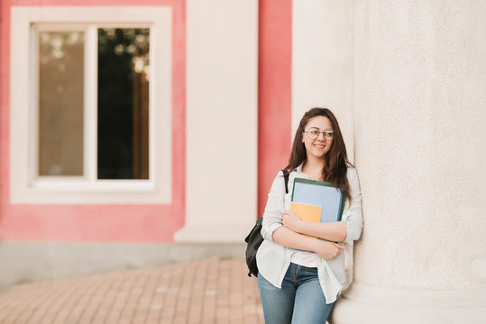 Portrait of a student girl with glasses. She stands in the background of a building with square windows as an area for text and holds notebooks and folders in her hands. The girl is preparing to enter