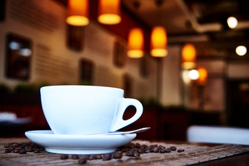 White cup with a spoon on a saucer on a bamboo napkin with scattered coffee beans.