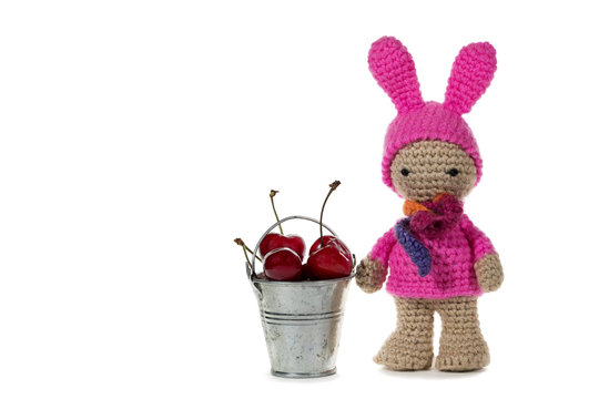 Dressed as a bunny crochet girl and a metal bucket with cherries, on white background. Amigurumi. Handmade