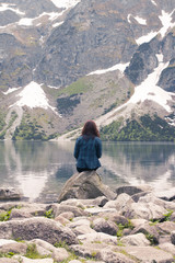 Young lonely brunette girl sitting on the shore of a blue lake in the mountains