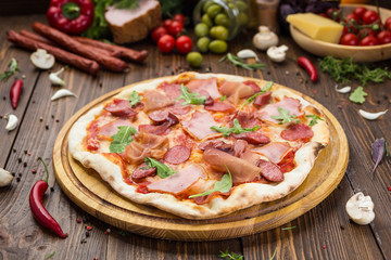 Delicious italian pizza with ham, bavarian sausages and prosciutto decorated by basil leaves on wooden background