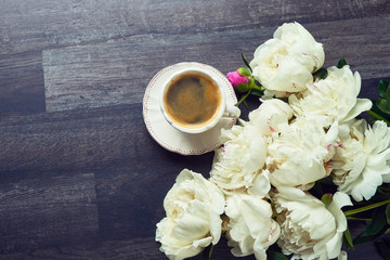 A cup of coffee and peony flowers on dark wooden background. Top view, copy space.
