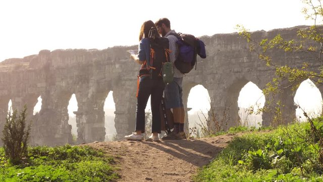 Young lovely couple backpackers tourists reading map pointing directions roman aqueduct arches in parco degli acquedotti park ruins in rome on romantic misty sunrise with guitar and sleeping bag slow
