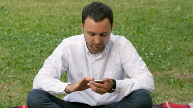 Handsome adult man sending a text message using cell phone at sunny evening outdoors in park