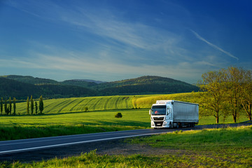 White truck driving on the asphalt road in a rural landscape with forested mountains in the...