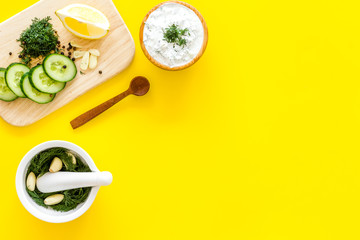 Preparing greek cucumber yogurt sauce. Bowl with yogurt near greenery, cucumber, oranges on cutting board on yellow kitchen desk top view copy space