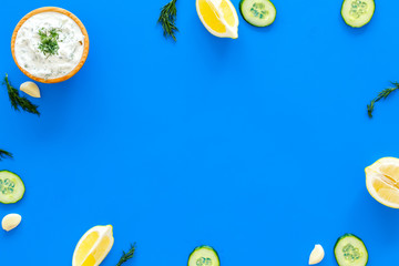 Preparing greek cucumber yogurt sauce. Bowl with yogurt near greenery, cucumber, oranges, garlic on blue kitchen desk top view copy space