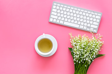 Cosy spring work desk concept. Computer keyboard, cup of tea and bouquet of lily of the valley flowers on pastel pink background top view copy space