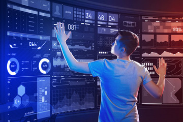 Working in the office. Smart professional IT manager thoughtfully looking at the transparent screen and touching the icons of it while working in his modern equipped office