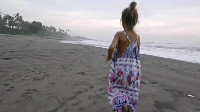 Following shot of little girl in summer dress running barefoot along ocean beach at sunrise