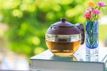 A teapot and moss rose flowers in blue vase on a book with nature background.