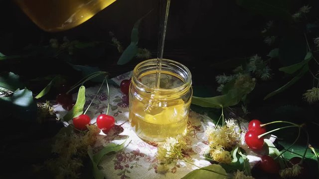 Pouring honey into glass jar, bunch of linden flowers and red cherry on wooden surface. Ray of sunlight. Dark rustic style.