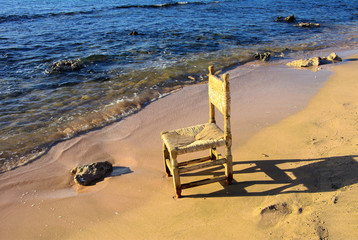 An old wicker chair from the vine is thrown out on an empty seashore against a background of yellow sand. Abandoned, unnecessary things. Vacation, tourism.