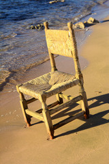 An old wicker chair from the vine is thrown out on an empty seashore against a background of yellow sand. Abandoned, unnecessary things. Vacation, tourism.