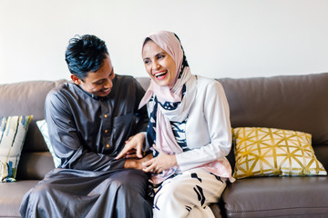 Fototapeta premium Portrait of a young Muslim couple in their home. They are dressed in traditional garb for Hari Raya (Ramadan) and sitting on a couch and tickling one another and laughing. 