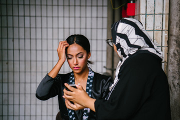 A portrait of two beautiful women taking a rest from a photo shoot. One wears a headscarf while the other fixes her hair. Both check the shots they've taken using a smartphone.