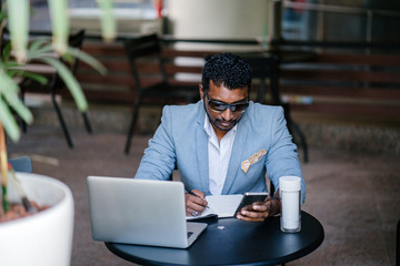 A cool Indian Asian businessman is working at a coworking space. He is looking at his smartphone while jotting down notes in his notebook.