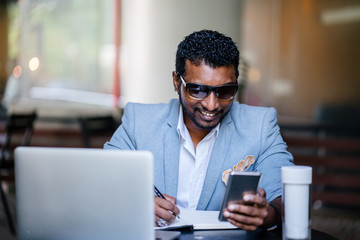 An Indian Asian man is sitting at a coffee shop and is glancing at his phone and writing in his notebook. His laptop is nearby and he is smiling as he does his work. He is dressed in a casual suit