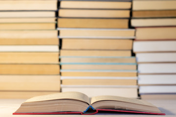 An open book in a blue hardback against the backdrop of many books stacked in piles
