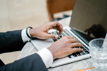 Close up shot of a man's hand as he types on a laptop keyboard. He has dark skin and a bracelet on...