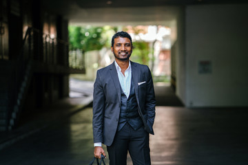 A stylishly dressed Indian Asian man stands in the street in the daytime. He is wearing a 3-piece suit with trendy shoes, a pocket square and earrings. He is holding a gym bag in one hand. 