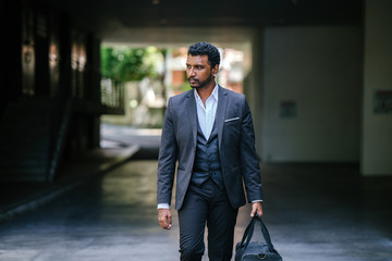 A stylishly dressed Indian Asian man is walking to go to the gym. He is wearing a 3-piece suit with a pocket square, and earrings. He is seen holding his gym bag in one hand.