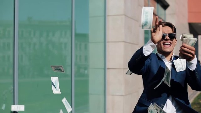 Young happy businessman scatter the dollars and funny dancing in the street. Money rain, falling dollars. It is located near the office center, slow motion. Successful business. Shot on Red Epic