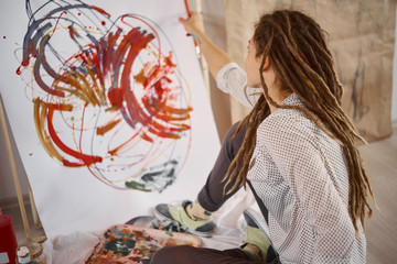 Girl painting abstract painting while sitting on studio floor.
