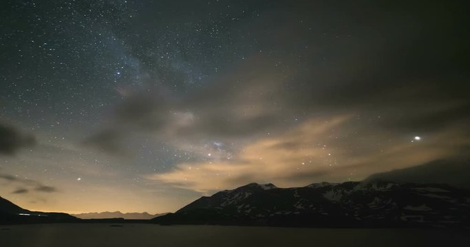Astro Time Lapse Milky way galaxy stars over the Alps, stormy sky, motion clouds, snowcapped mountain range and lake.