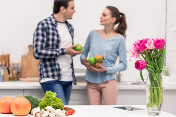 healthy food lying on table at kitchen with case of flowers while beautiful adult couple chatting blurred on background