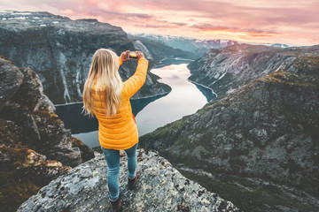 Woman taking photo by smartphone on mountain cliff over lake traveling in Norway adventure...