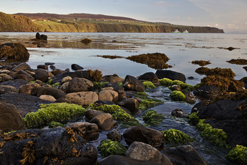 Pebble beach with boulders covered in seaweed in the foreground and steep sea cliffs in the distance