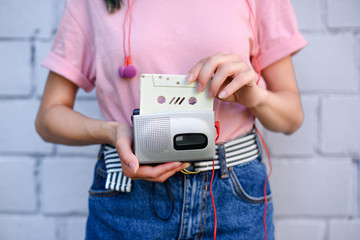 cropped shot of woman with earphones holding retro cassette player and audio cassette in hands against white brick wall