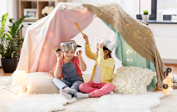 childhood and hygge concept - happy little girls with cooking pots playing in kids tent at home