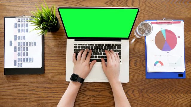 Male hands typing on laptop with green screen on a desk with clipboards on it. Top view footage