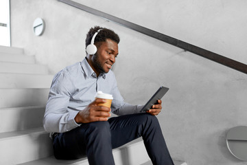 business, people and technology concept - african american businessman with headphones and tablet pc computer listening to music at office coffee break