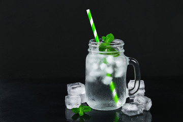 Ice water with ice and a mint leaf and a straw in a mason jar on a black background.