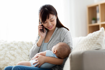 Obraz na płótnie Canvas family, technology and motherhood concept - happy smiling young asian mother with sleeping baby calling on smartphone at home