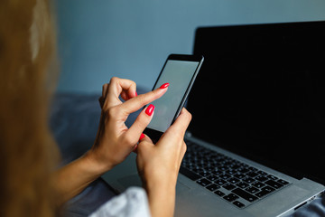 Lovely blonde woman phoning with her smartphone lying on her bed next to her notebook. Mobile phone with blank screen in female hands. Сoncept of chating, mobile banking, online payment.